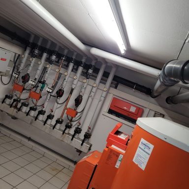 Heizungsraum mit Rohren, Pumpen und einem orangefarbenen Heizkessel.