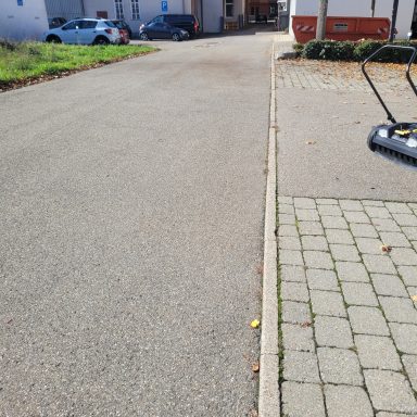 Straße mit Fußweg, links ein Rasenmäher, im Hintergrund Gebäude und Autos.