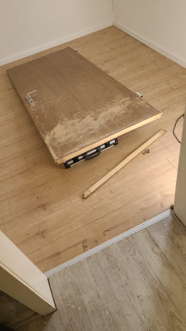 Ein Holzbrett liegt auf einem hellen Boden, daneben liegt ein weiterer Holzstab.